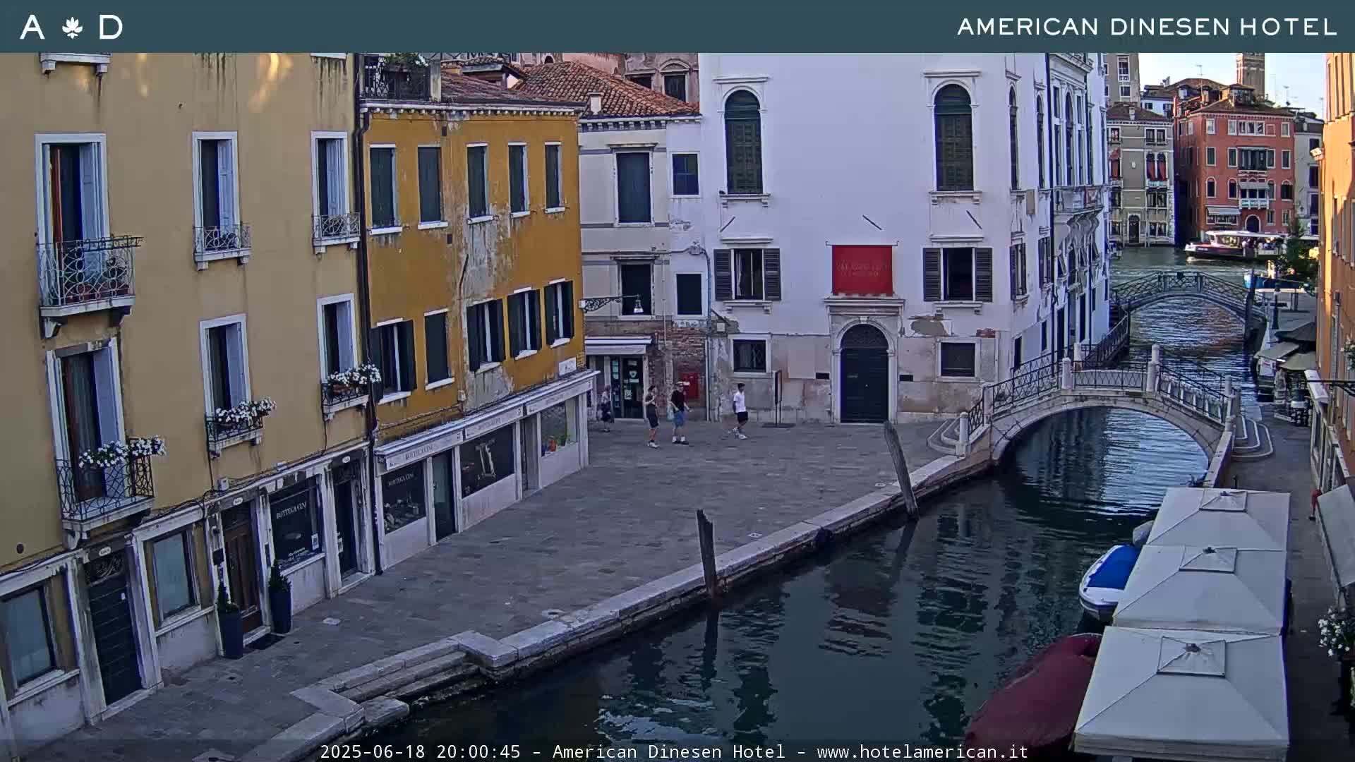 A tranquil canal scene in Venice, Italy, shows several buildings lining the waterway, a small bridge, and a few people walking along the canal under a clear evening sky.