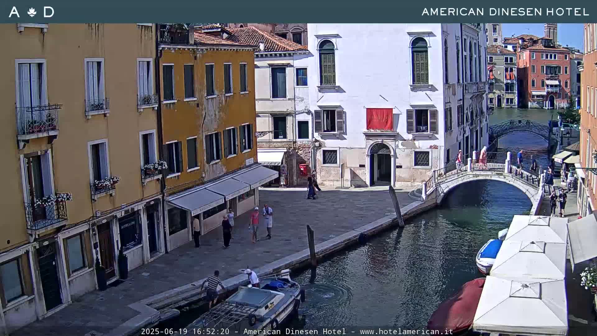 A sunny Venetian canal scene shows a bridge, several buildings, a few people walking, and a small boat near a waterfront.