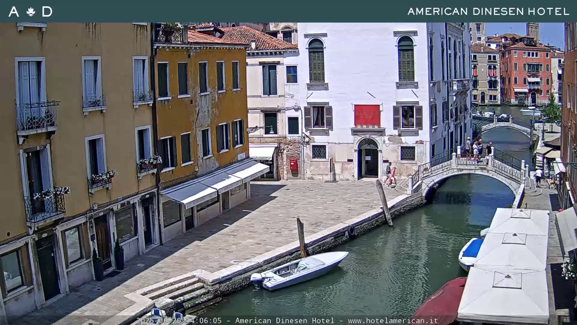 A sunny day in Venice shows a canal with a small motorboat and a pedestrian bridge, flanked by buildings with awnings and balconies.