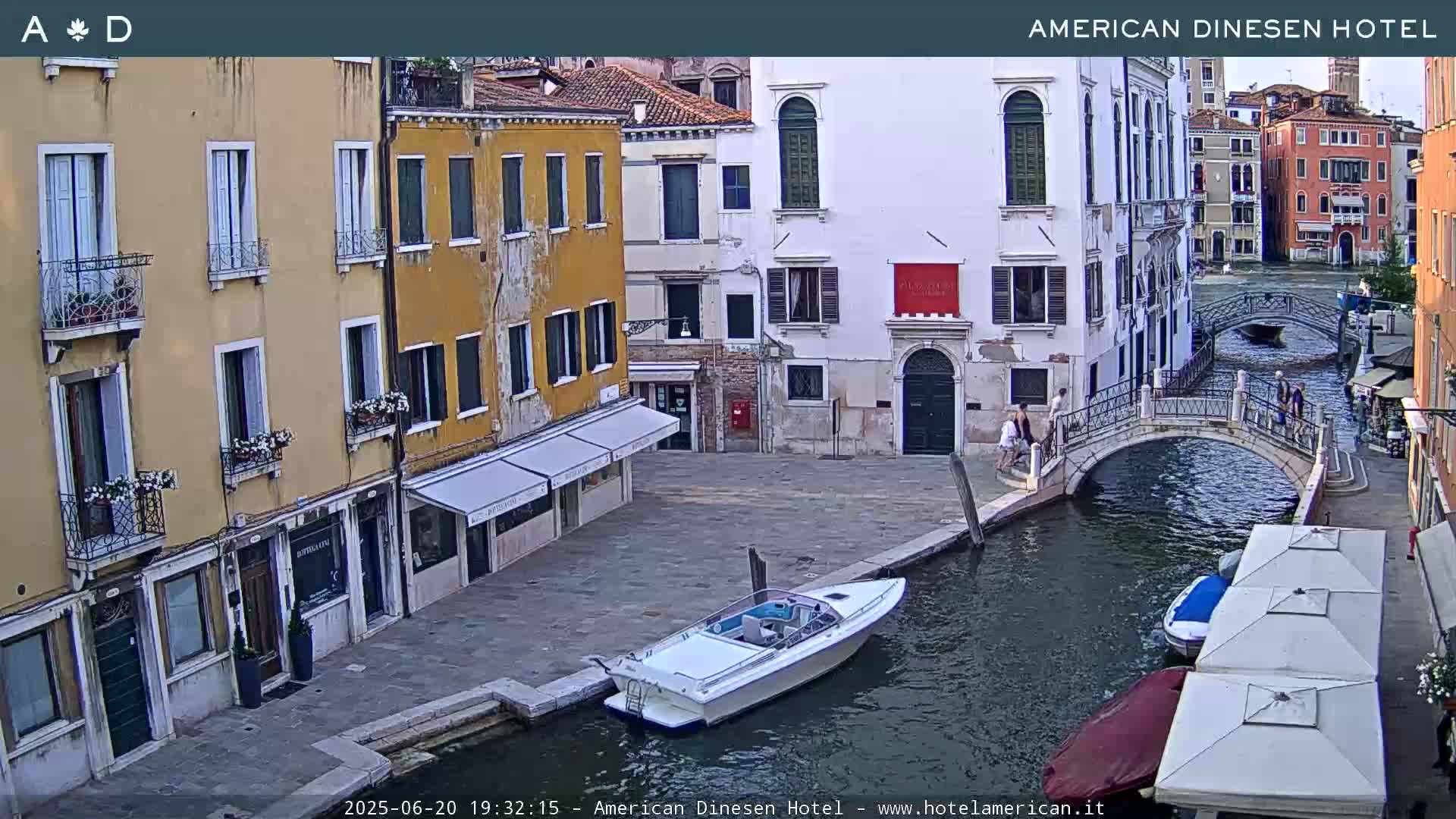 A canal in Venice, Italy, with several buildings lining its banks, a small motorboat docked, and a footbridge spanning the waterway, under a partly cloudy sky.