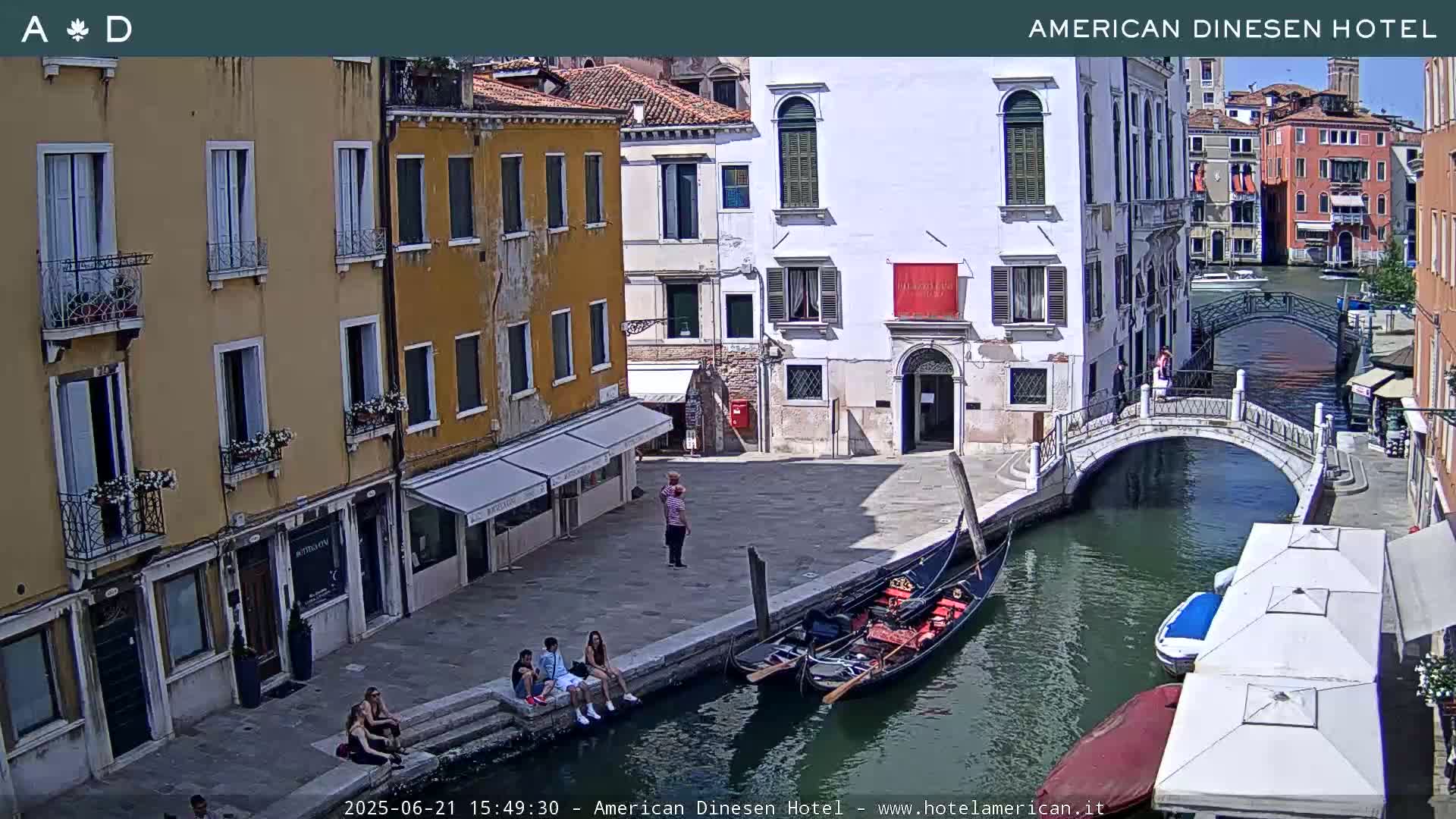 A sunny Venetian canal scene shows several buildings, a gondola, a small bridge, and a few people sitting and standing on the canal's edge.