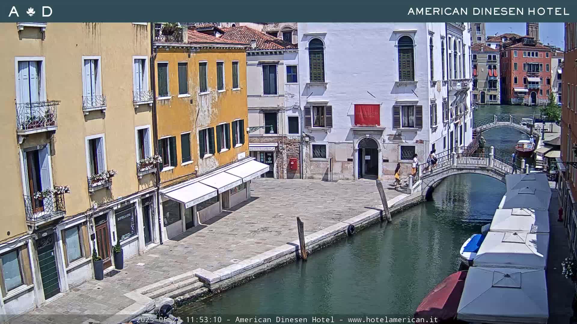 A sunny Venetian canal scene shows several buildings, a pedestrian bridge, and small boats moored along the waterway.