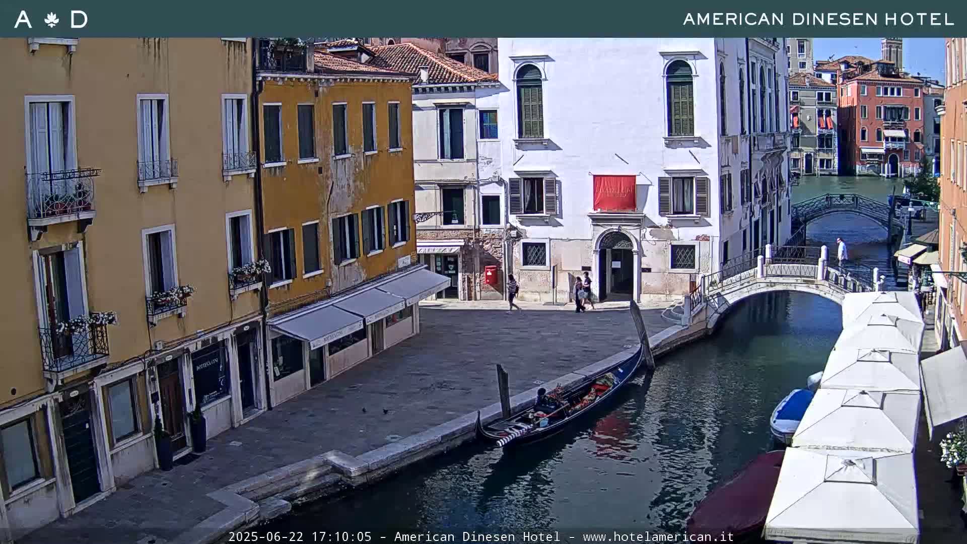 A gondola is moored along a canal in Venice, Italy, on a sunny day, with several buildings lining the waterway and a small bridge crossing the canal.