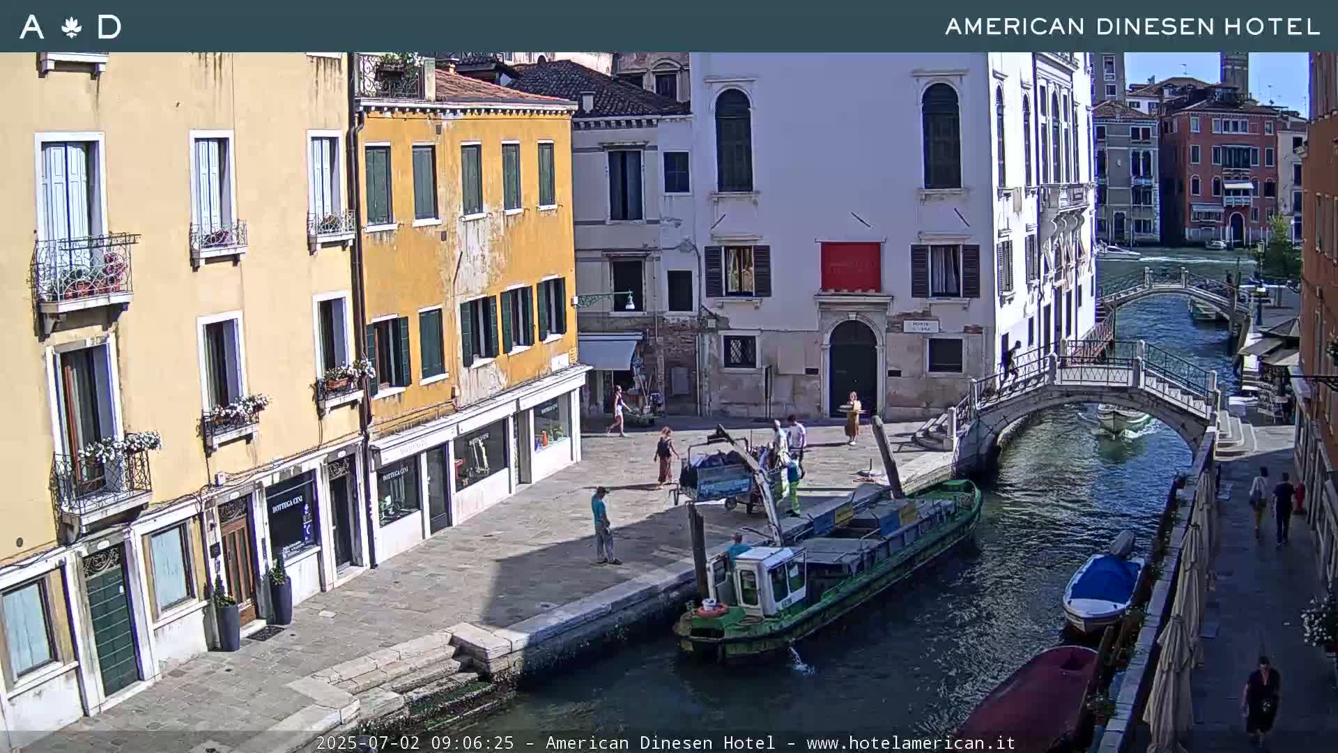 A sunny day in Venice shows a canal with boats, a pedestrian bridge, and several buildings lining the waterway, with people walking along the waterfront.