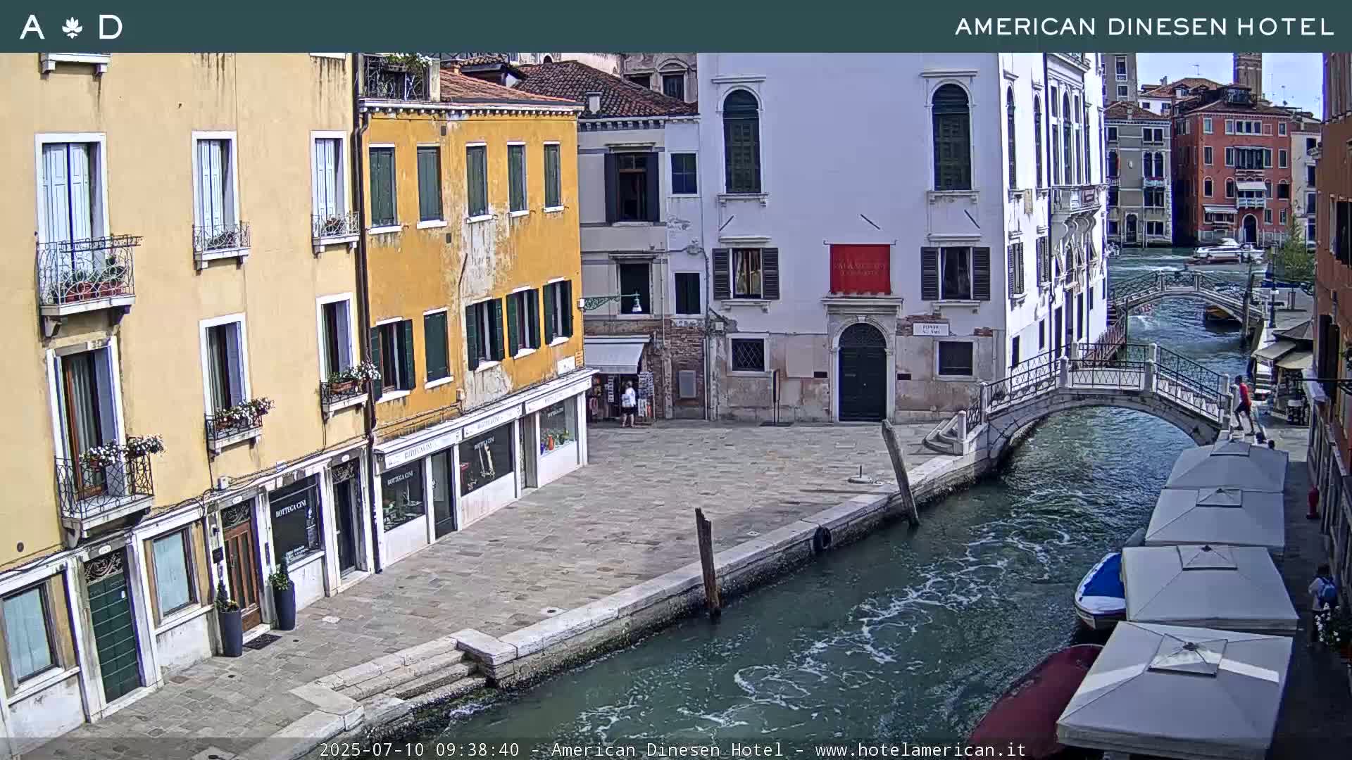 A sunny Venetian canal scene shows colorful buildings lining the waterway, a small footbridge, and several outdoor patio umbrellas.