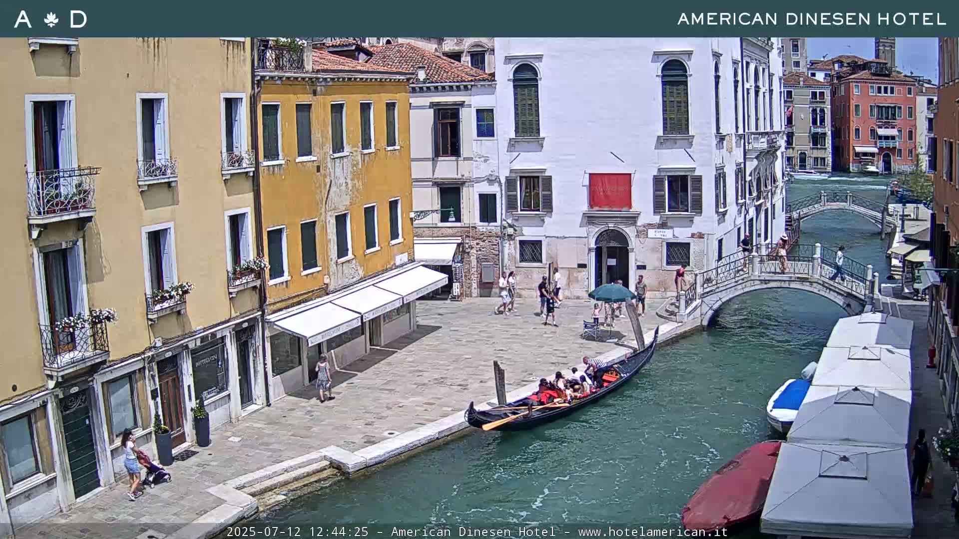 A sunny Venetian canal scene shows colorful buildings lining the waterway, a small footbridge, and several outdoor patio umbrellas.