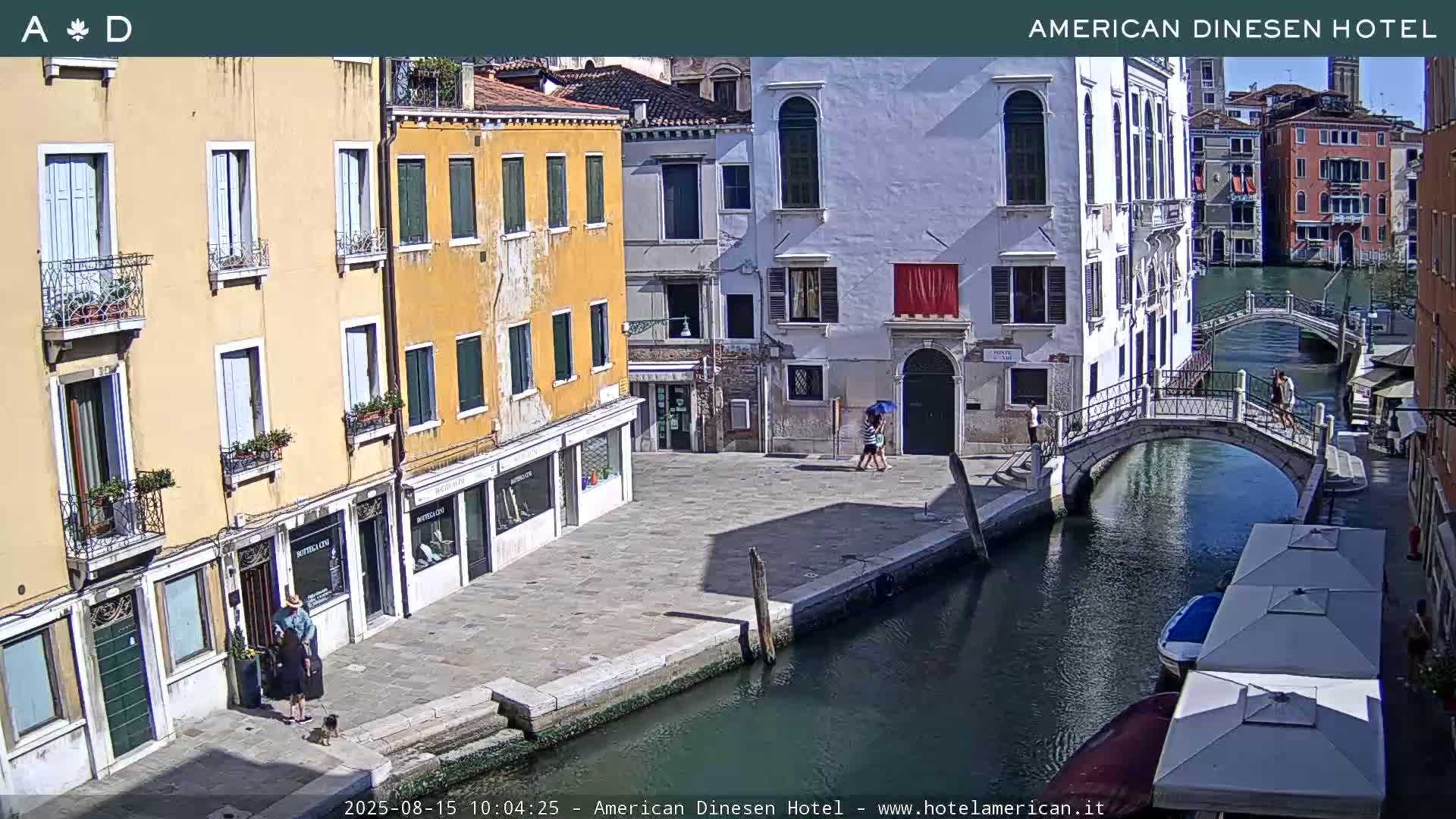 A sunny day in Venice shows a canal with a small bridge, several people walking, and buildings lining the waterway.