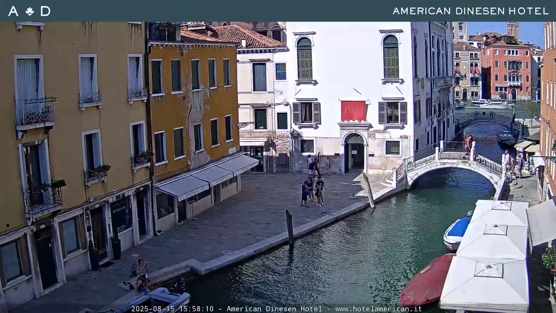 A sunny day in Venice shows a canal with a small bridge, several people walking, and buildings lining the waterway.