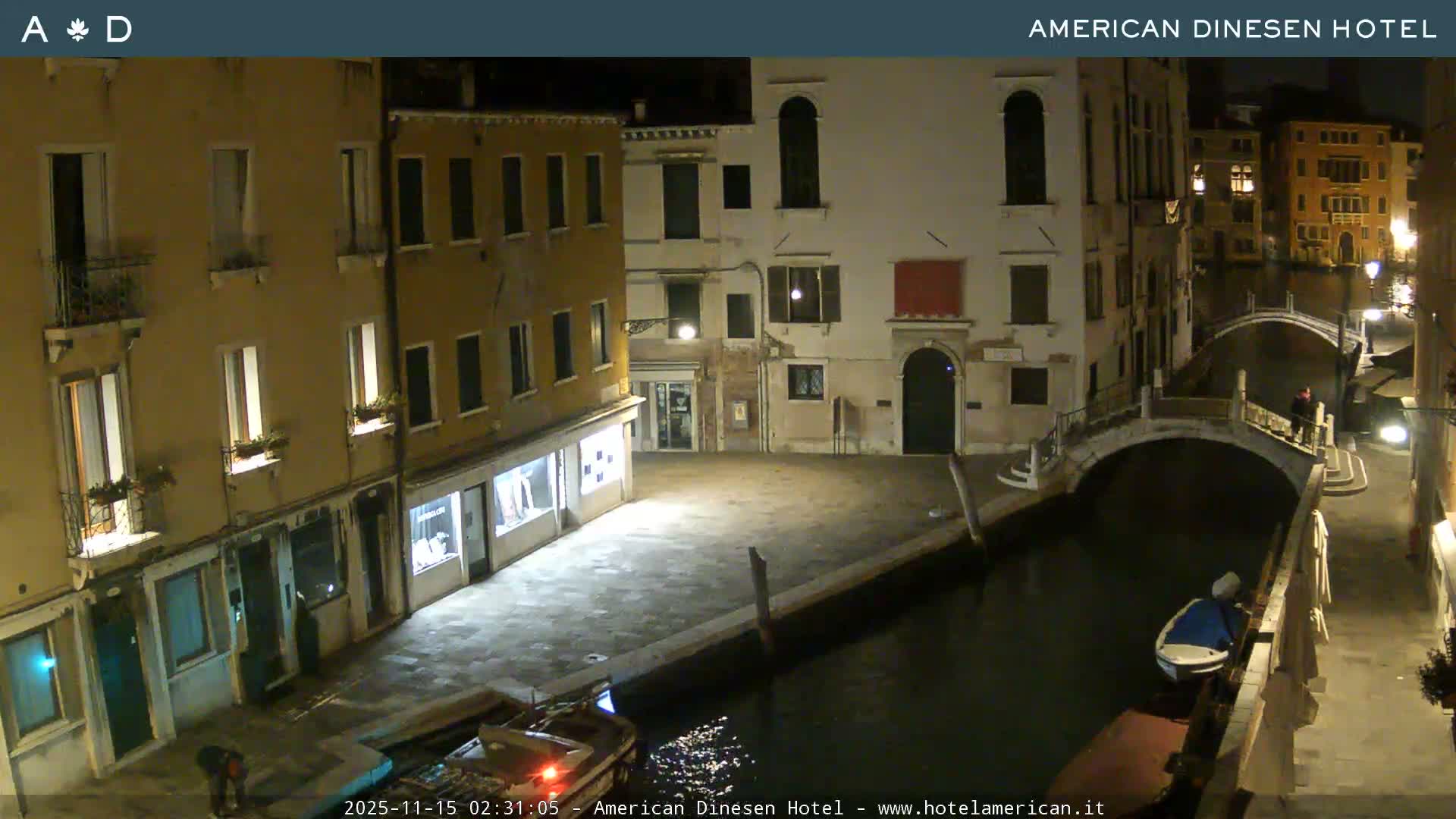 A tranquil night scene in Venice shows illuminated historic buildings lining dark canals, traversed by arched bridges, with a few moored boats and individuals visible under a clear, dark sky.