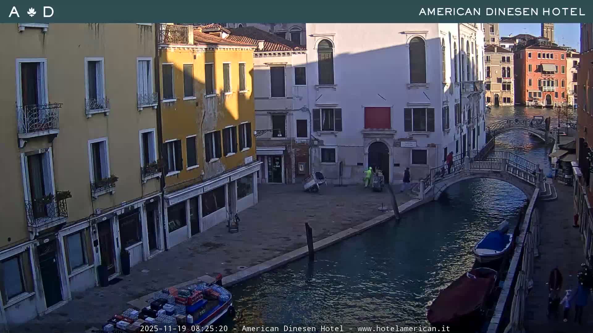 A tranquil night scene in Venice shows illuminated historic buildings lining dark canals, traversed by arched bridges, with a few moored boats and individuals visible under a clear, dark sky.