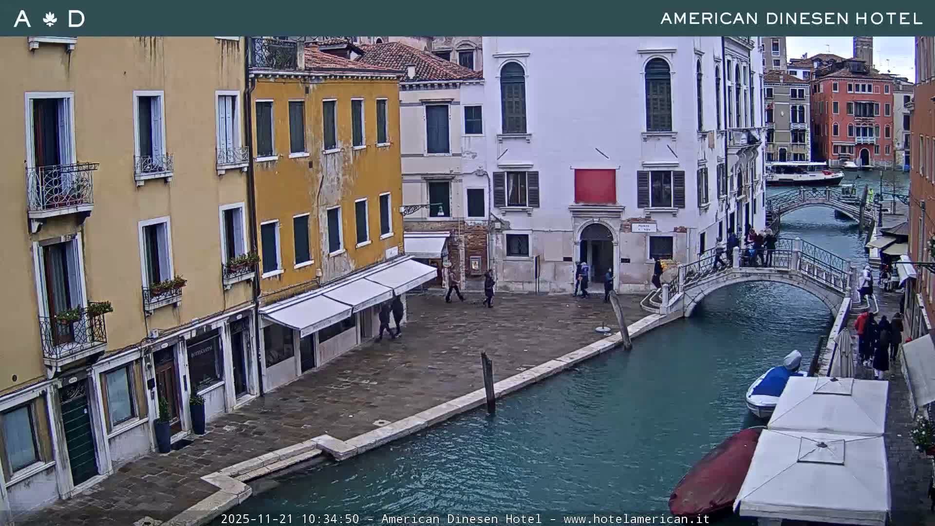 An overcast day in Venice reveals a narrow canal lined with traditional multi-colored buildings, spanned by stone arched bridges carrying pedestrians, and dotted with several moored boats.