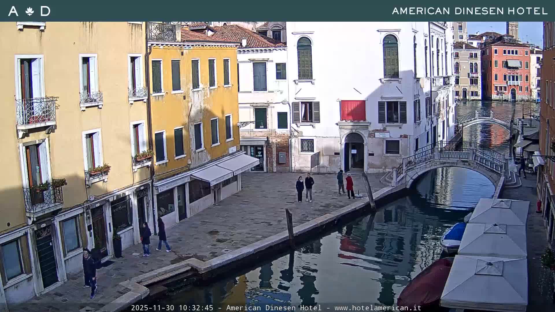 On a clear and sunny day, a vibrant Venetian scene features historic, colorful buildings lining a canal traversed by a stone bridge, with several people walking on the adjacent paved square and boats gently floating on the water.