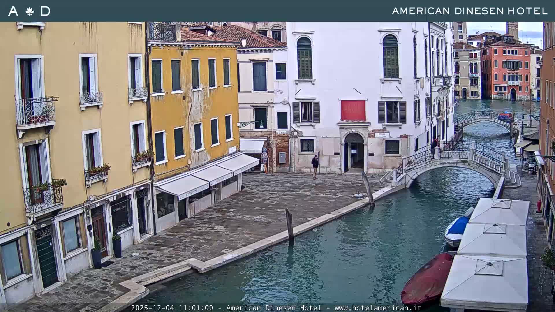 A scenic view of a Venetian canal on an overcast day features colorful historic buildings lining a waterway with several boats, two arched bridges, and a few pedestrians.