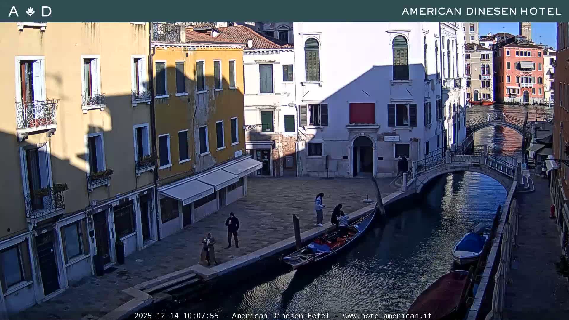 A scenic view of a Venetian canal on an overcast day features colorful historic buildings lining a waterway with several boats, two arched bridges, and a few pedestrians.