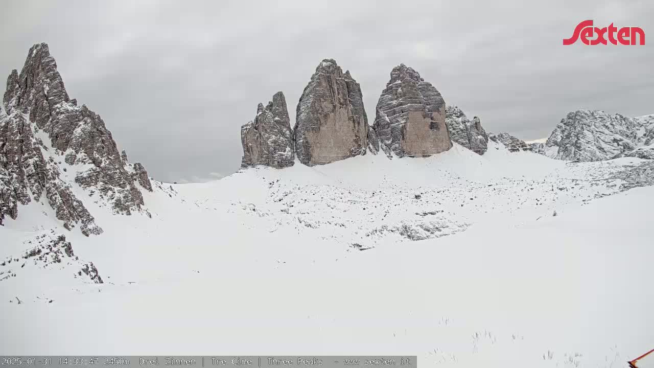 A snow-covered landscape features a prominent group of three tall, rocky peaks under an overcast sky.