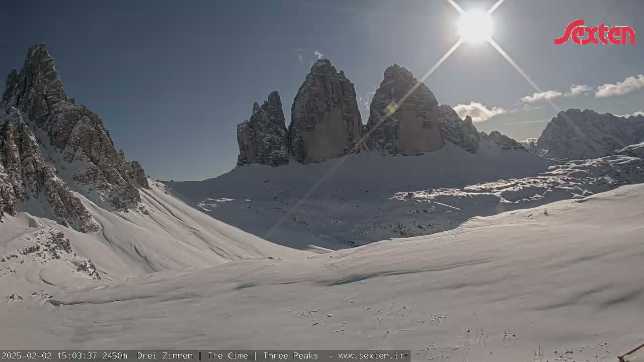 A sunny, snow-covered mountain landscape features a prominent group of jagged peaks under a bright sun.