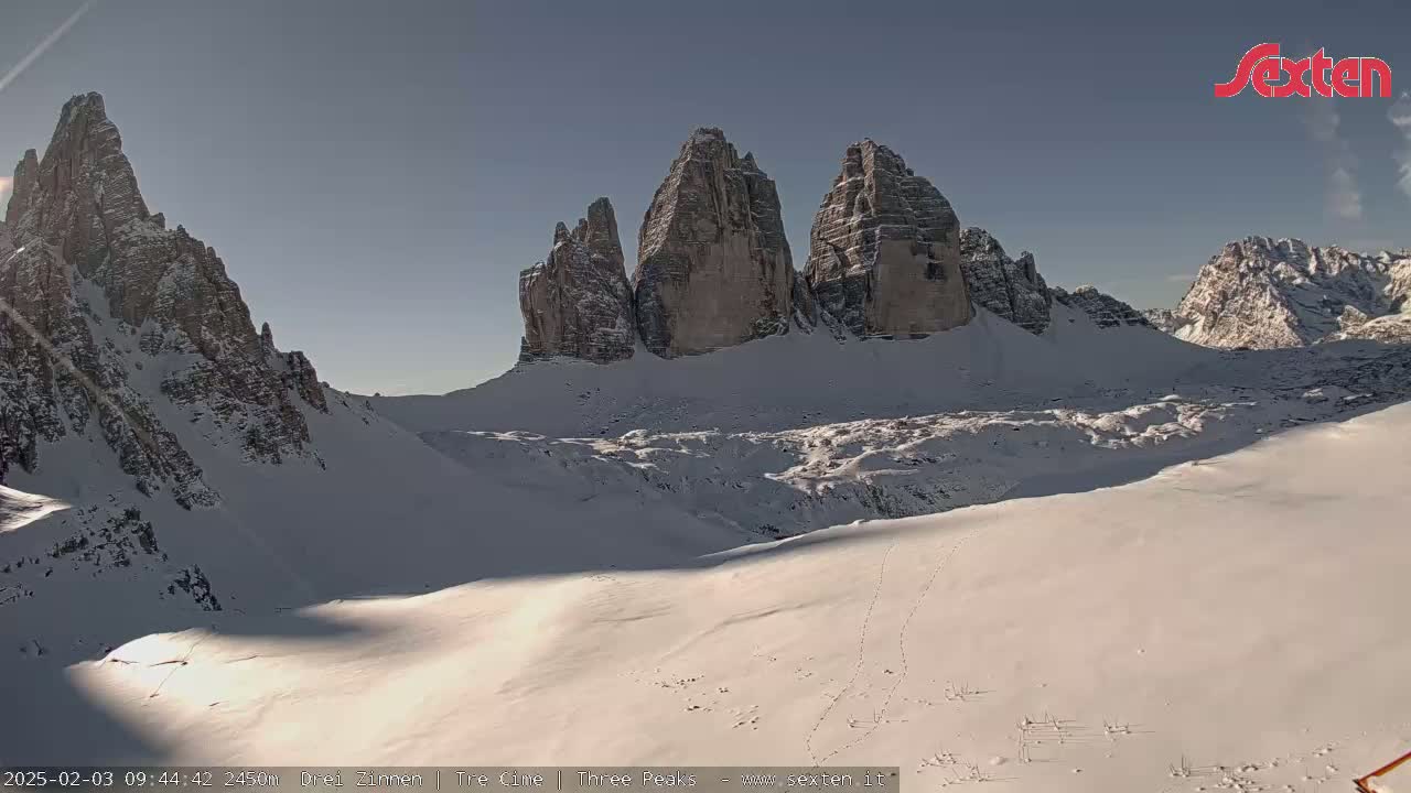 A snow-covered valley is viewed from a high vantage point, showcasing several prominent rock formations under a clear sky.