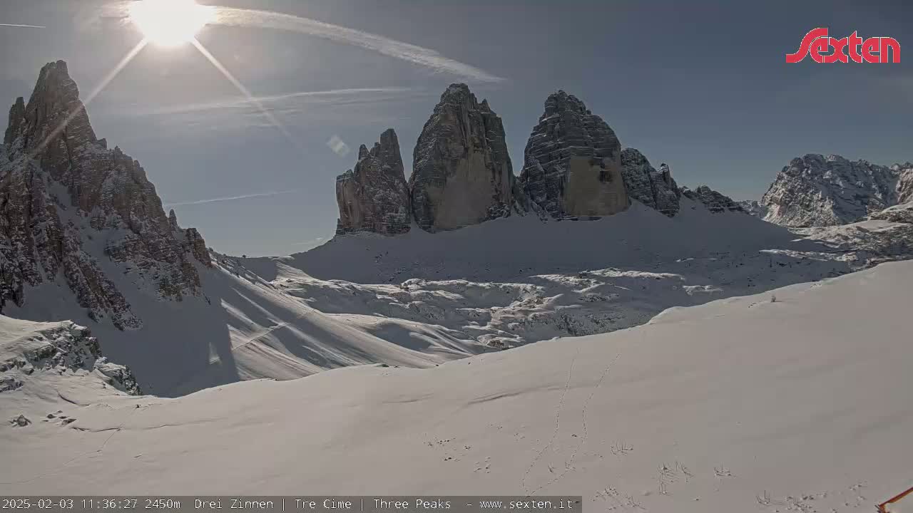 A sunny, snow-covered mountain landscape features a prominent rock formation of three tall peaks.