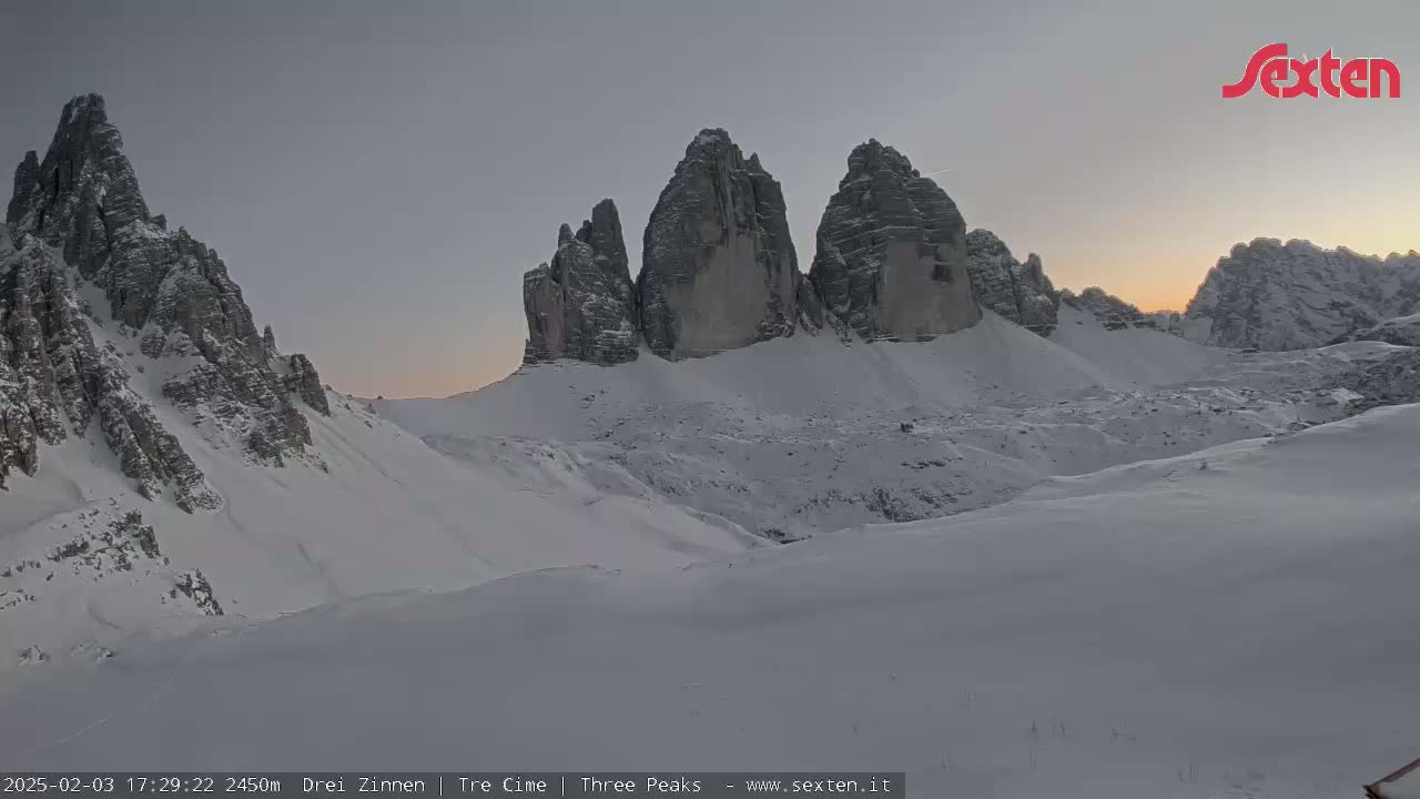 A snow-covered mountain range with several prominent peaks is illuminated by the soft light of sunrise or sunset under a pale sky.