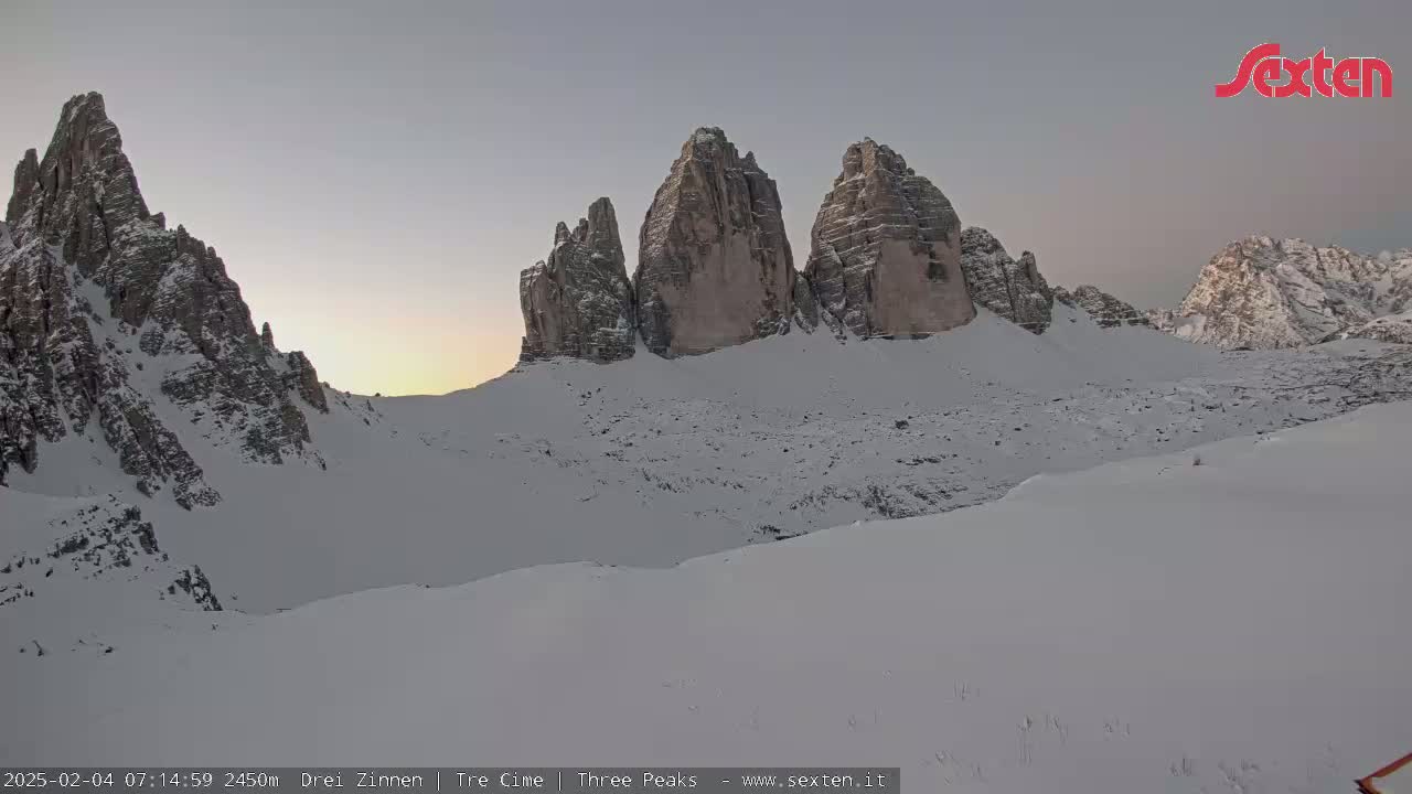 A snow-covered mountain range with several prominent peaks is visible at sunrise under a clear, pale sky.