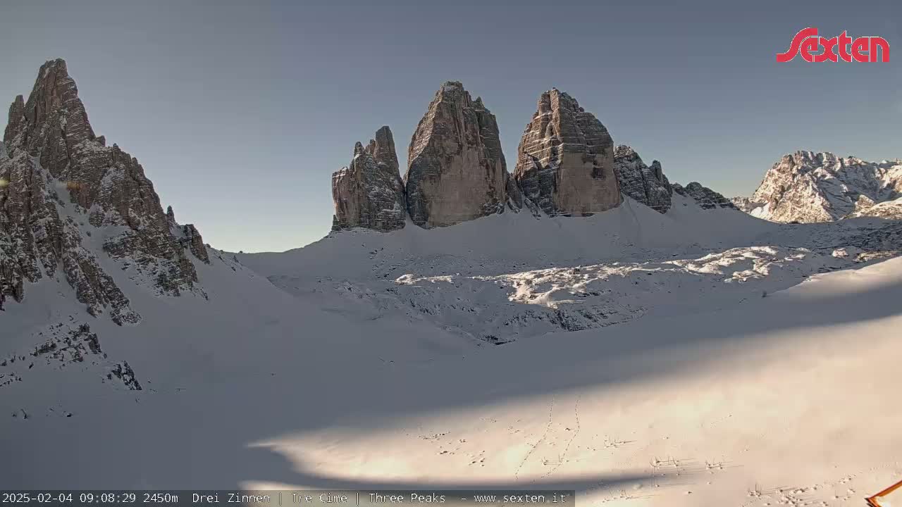 A snow-covered mountain valley with prominent, snow-dusted rock formations in the background under a clear, bright sky.