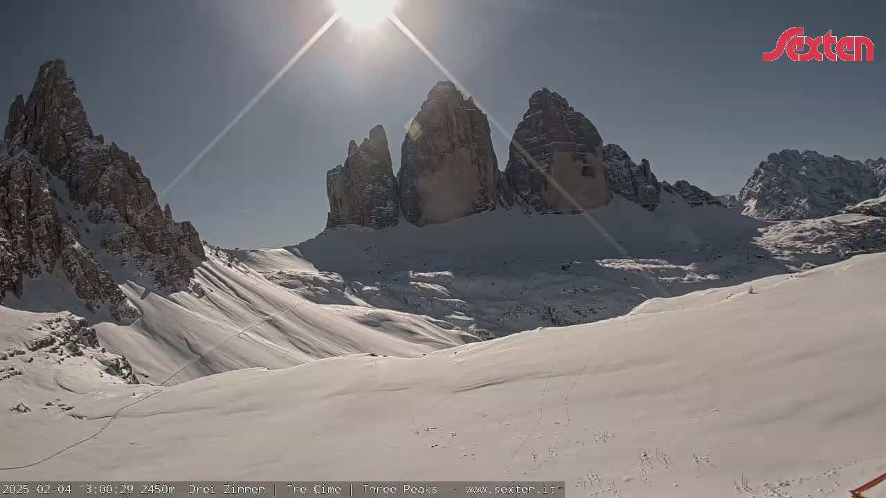 A sunny, snow-covered mountain landscape features several prominent rock formations in the background.