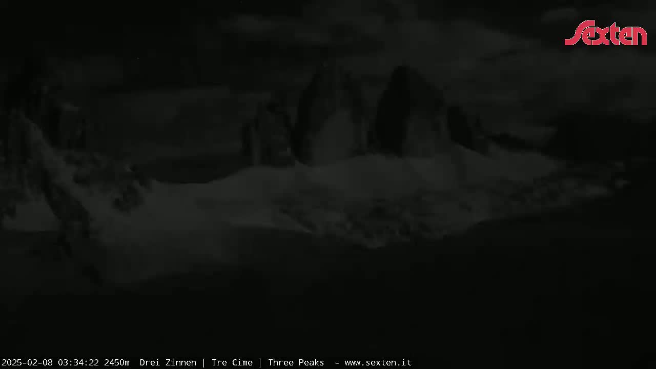A dark, nighttime image shows snow-covered, indistinct mountain peaks under a cloudy sky.