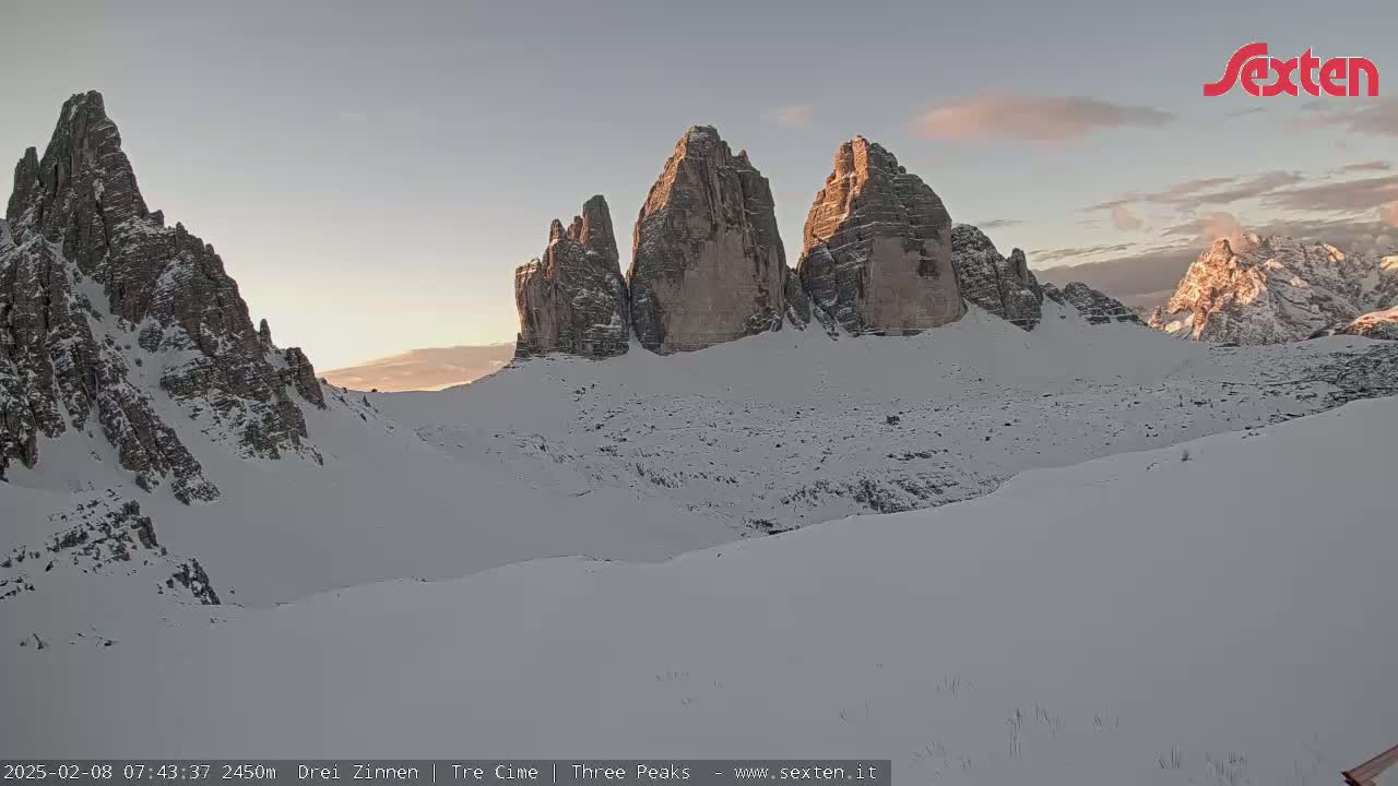A snow-covered mountain range, with several prominent peaks, is illuminated by the soft light of sunrise or sunset.