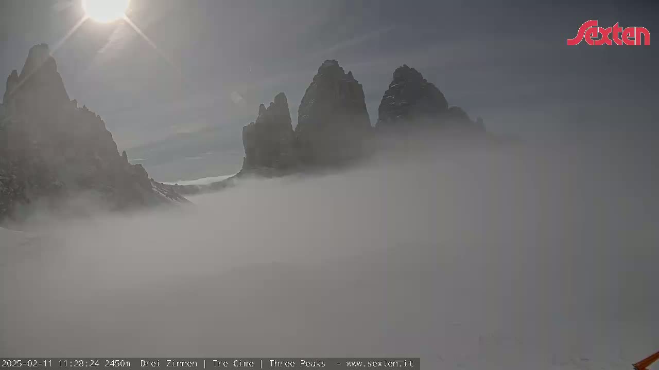 Three rocky peaks are partially obscured by a thick fog under a bright, sunny sky.