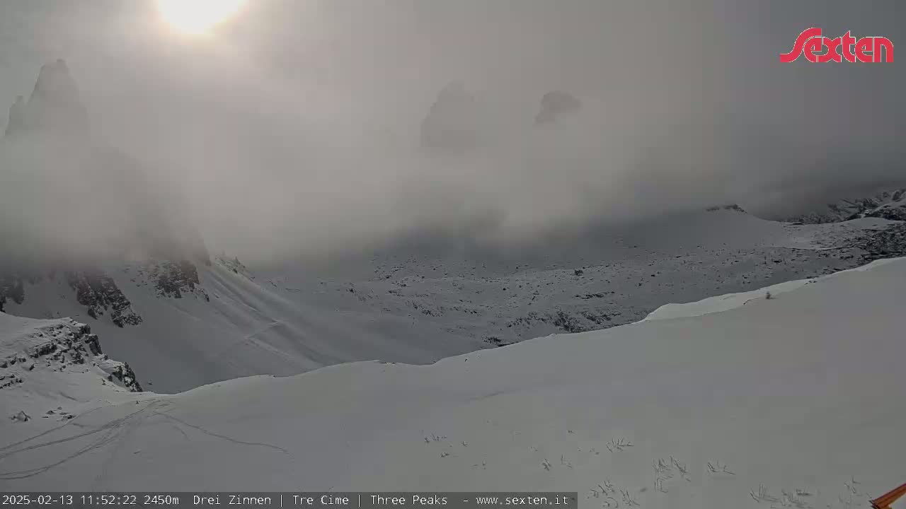 A snow-covered mountain landscape is partially obscured by fog, with a bright sun visible through the haze.