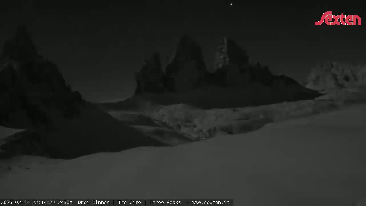 A nighttime, snow-covered landscape features dark, jagged mountain peaks under a clear, starry sky.