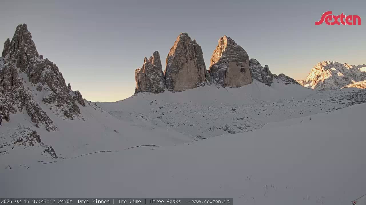 A snow-covered mountain range, illuminated by the morning sun, features prominent, sharply peaked rock formations in the center.