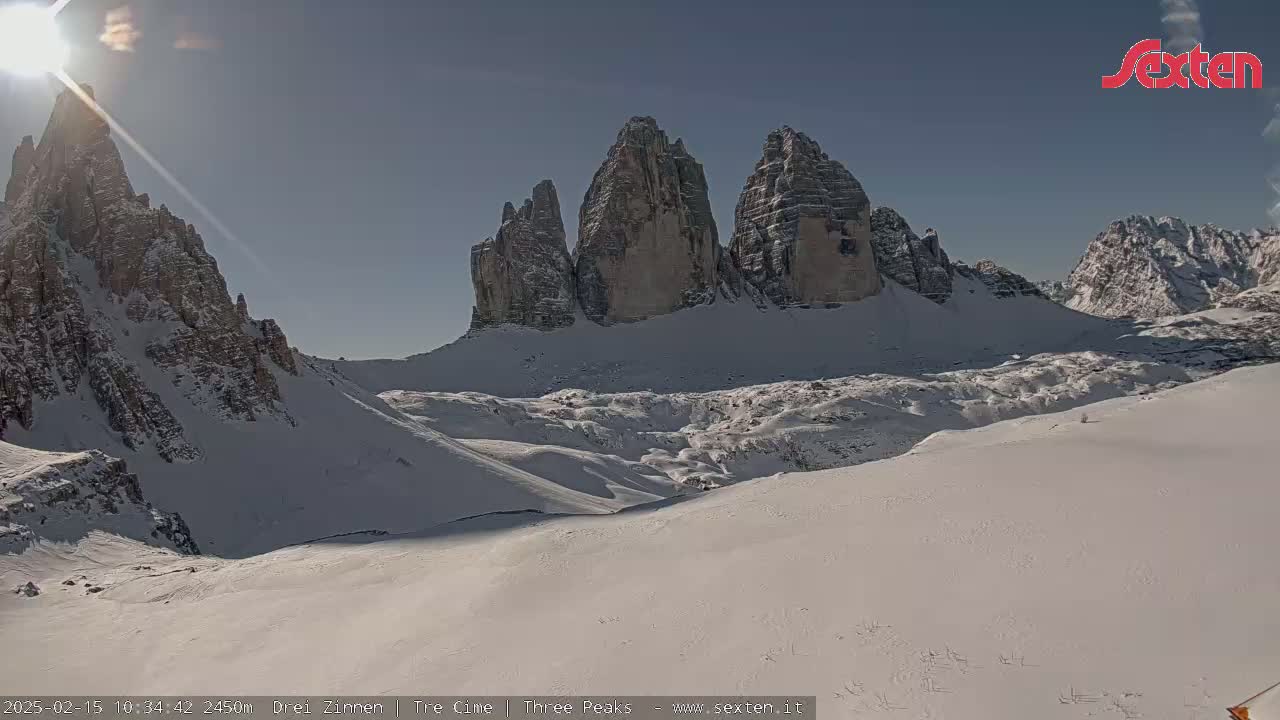 A sunny, snow-covered mountain landscape features a prominent group of tall, rocky peaks under a clear blue sky.