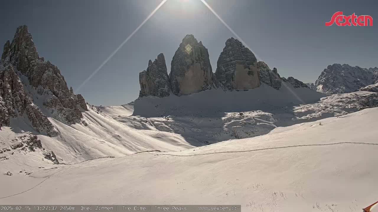 A sunny, snow-covered mountain landscape features a prominent group of three peaks rising above a valley.