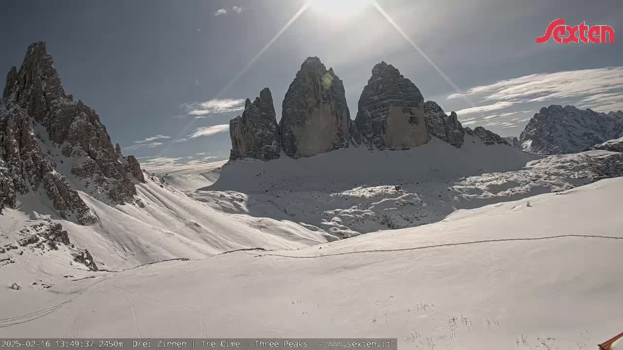 A sunny, snow-covered mountain landscape features several prominent rock formations under a mostly clear sky.