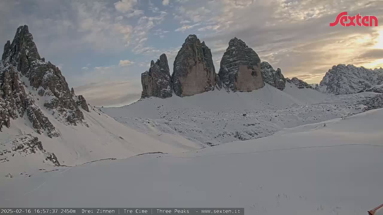 A snow-covered mountain landscape at sunset, featuring several prominent rock formations.