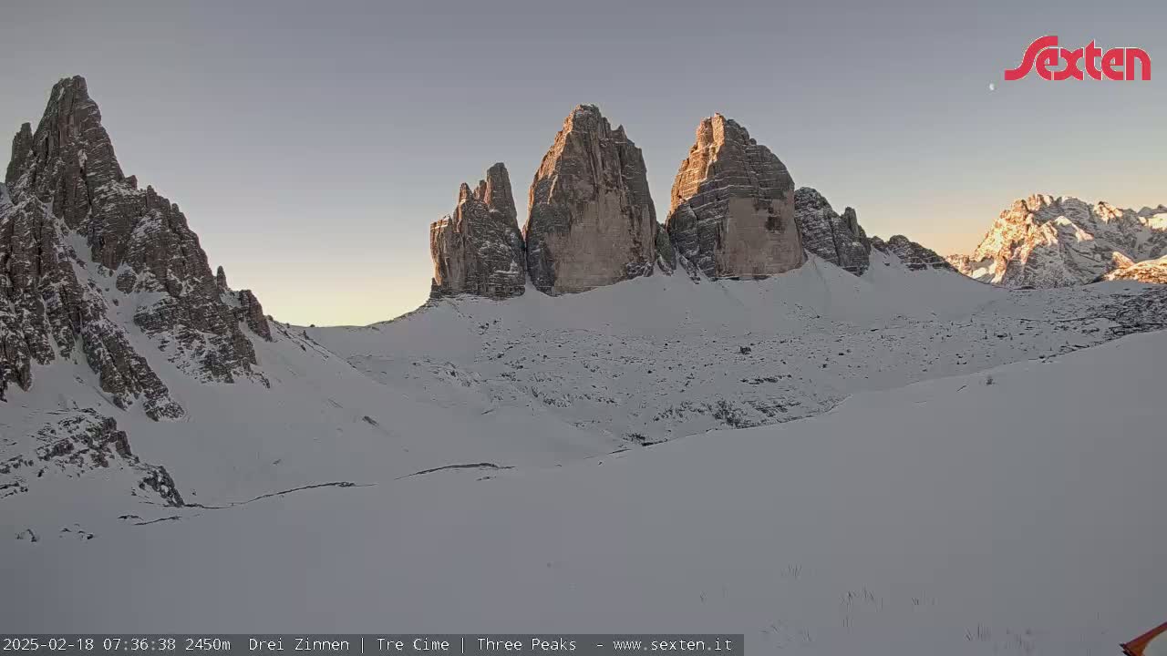 A snow-covered mountain valley at sunrise, featuring a prominent group of three tall, rocky peaks.
