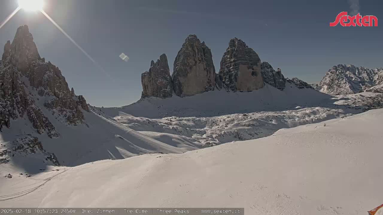 A sunny, snow-covered mountain landscape features a prominent group of three tall, rocky peaks in the background.