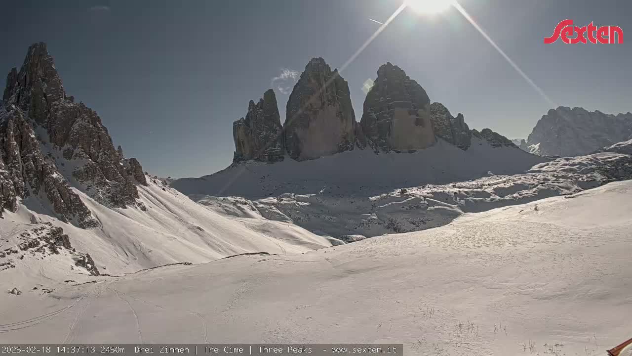 A sunny, snow-covered mountain valley is visible, with tall, rocky peaks rising in the background.