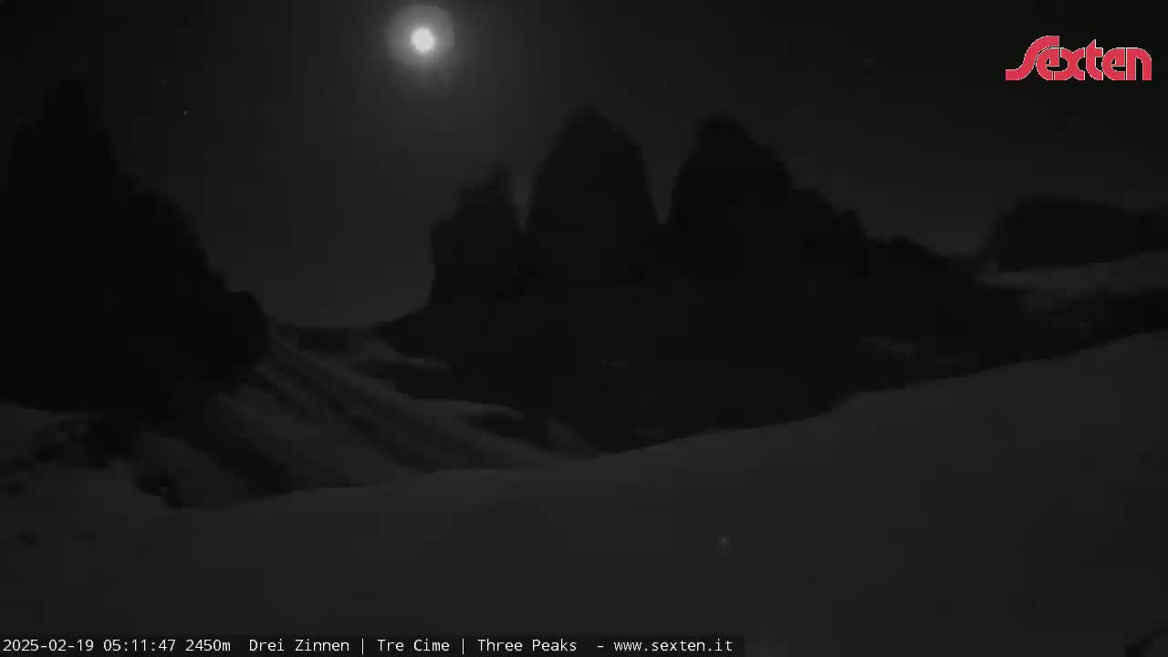 A bright moon illuminates snow-covered ground and three dark, tall mountain peaks under a clear night sky.