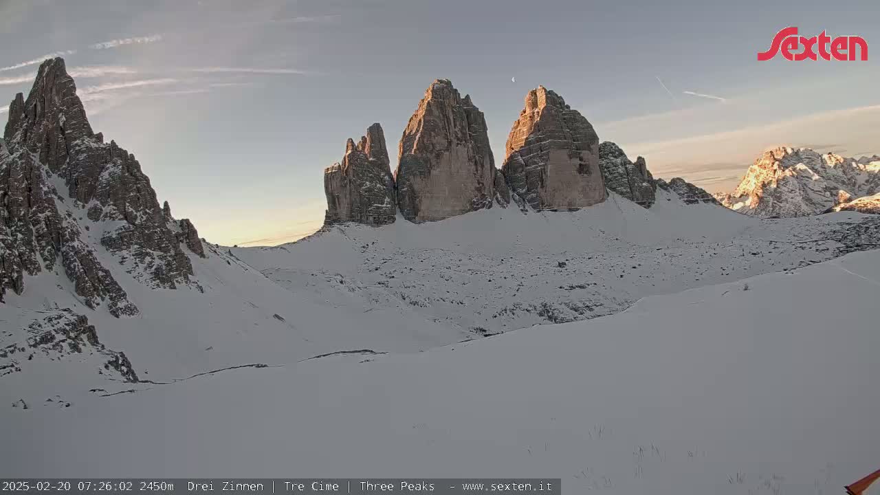 A snow-covered mountain range, featuring several prominent peaks, is bathed in the soft light of sunrise or sunset under a clear sky.