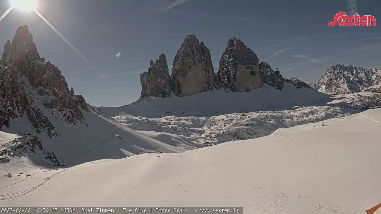 A sunny, snow-covered mountain range with several prominent rock formations is visible under a bright, mostly clear sky.