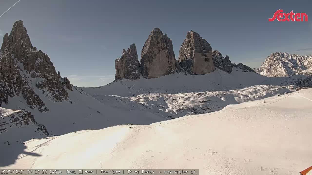 A sunny, snow-covered mountain landscape features a prominent group of jagged peaks in the background.