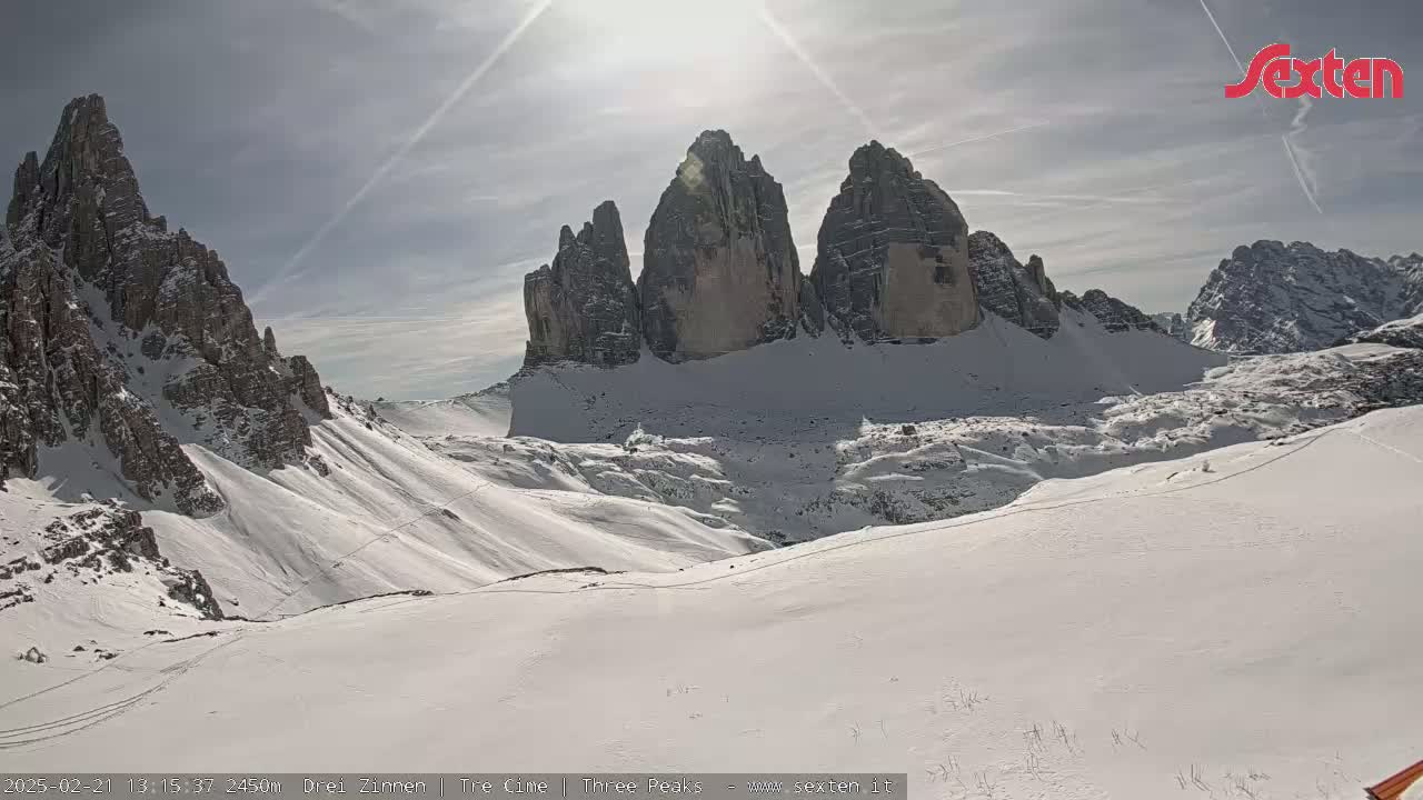 A sunny, snow-covered mountain landscape features several tall, jagged peaks under a mostly clear sky.