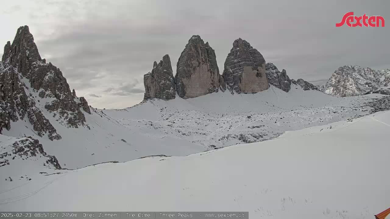 A snow-covered mountain valley is seen with several tall, rocky peaks in the background under an overcast sky.