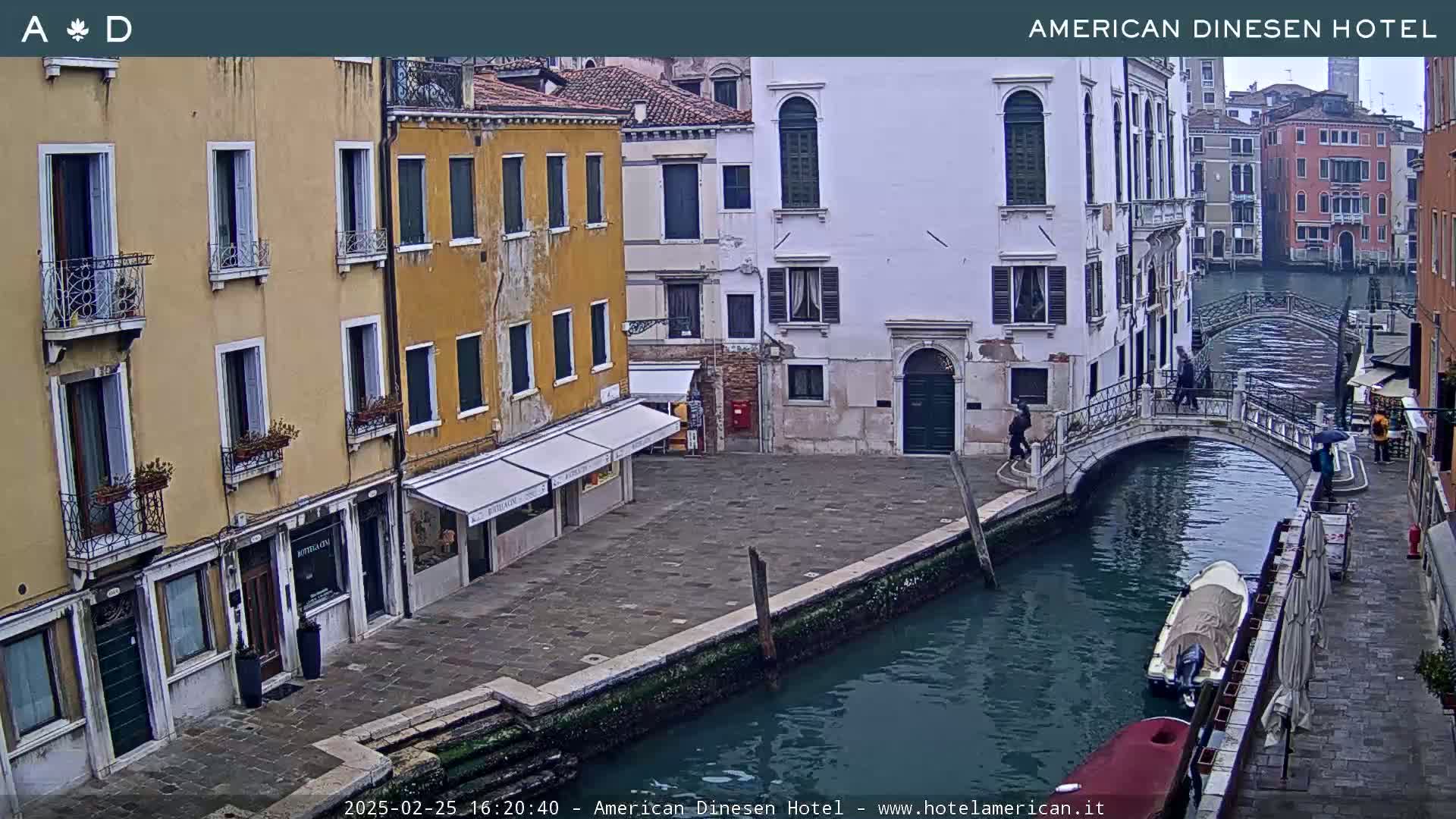 A view of a canal in Venice on an overcast day, showing colorful buildings, a stone bridge, and several small boats.