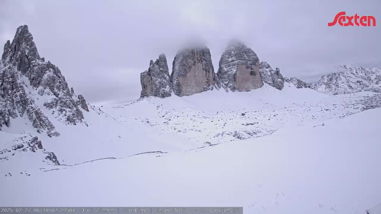A snow-covered valley with three large rock formations partially obscured by fog in the background under an overcast sky.