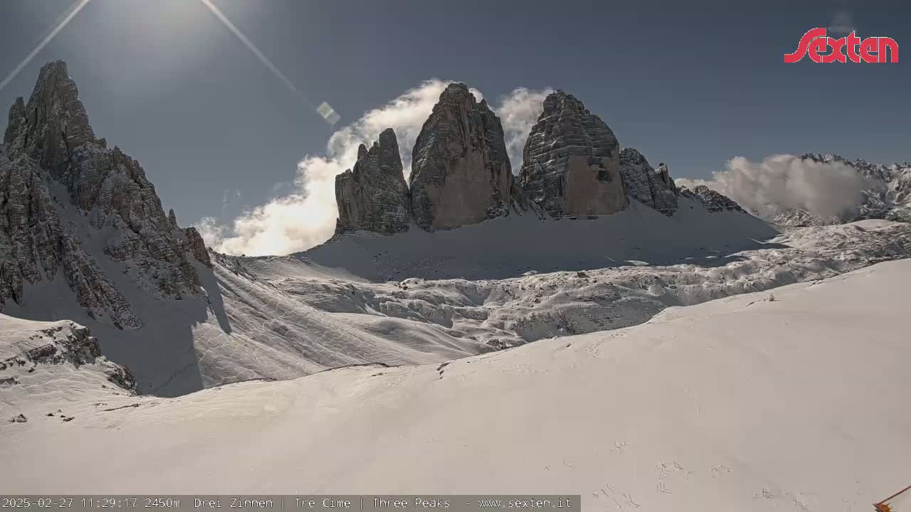A sunny, snowy mountain landscape features three prominent rock formations partially obscured by clouds.