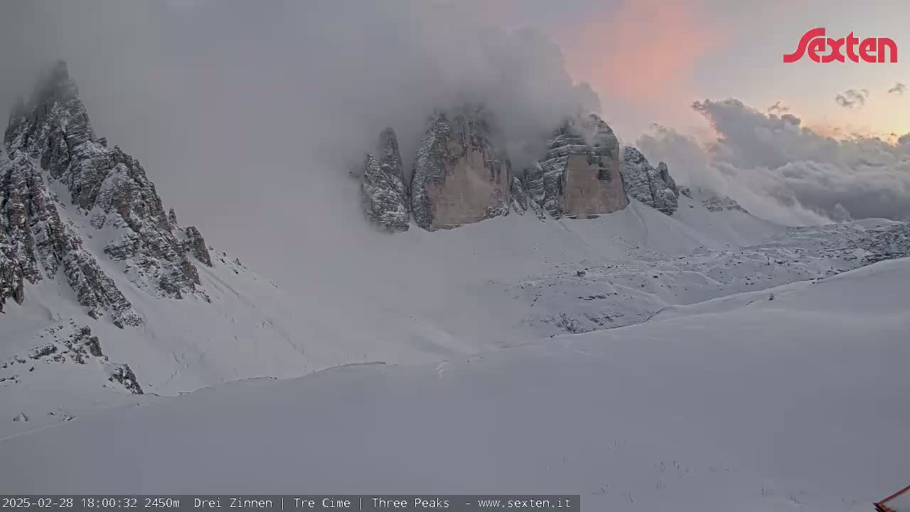 Snow-covered mountains partially obscured by clouds and fog under a pinkish sky.