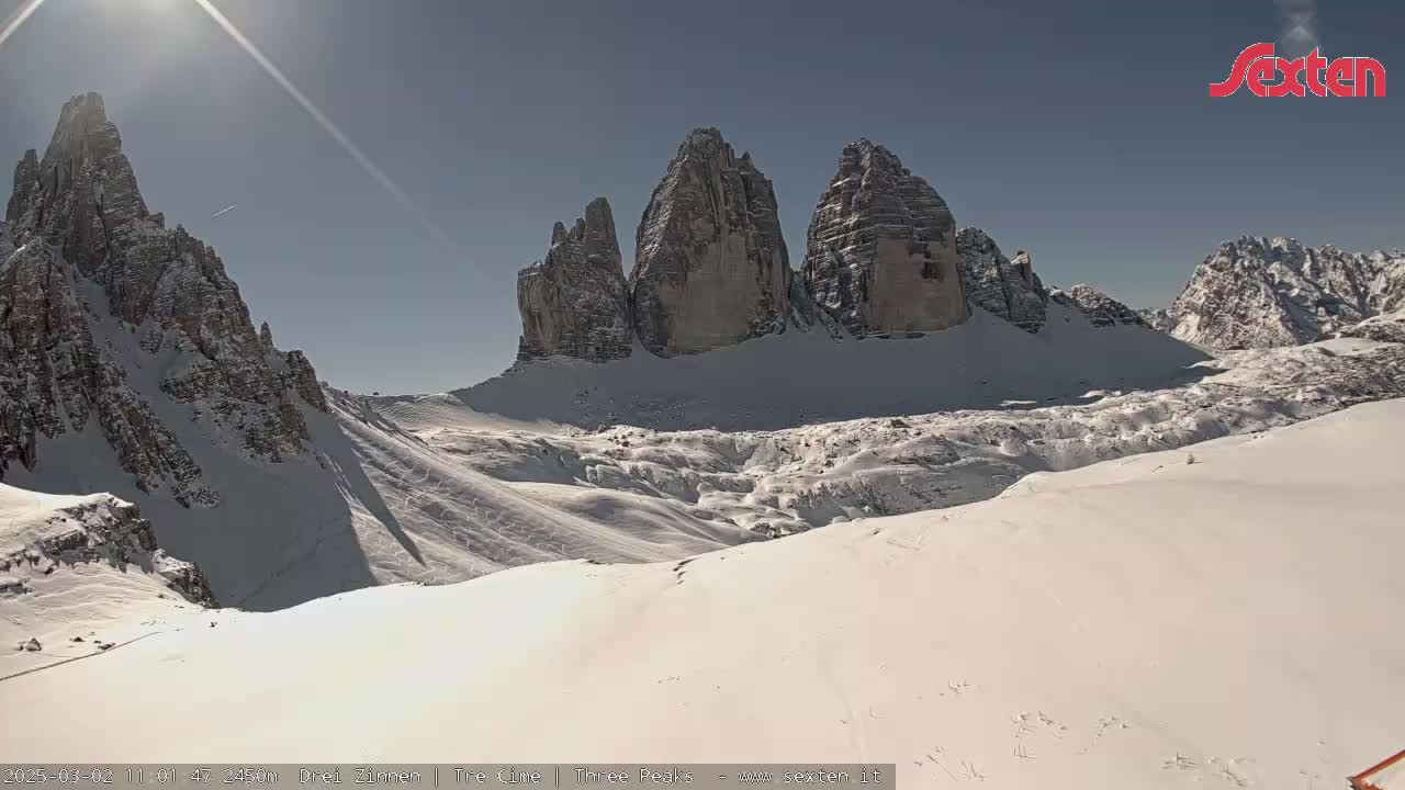 A sunny day reveals snow-covered mountains, with prominent rock formations rising from a valley filled with snow.
